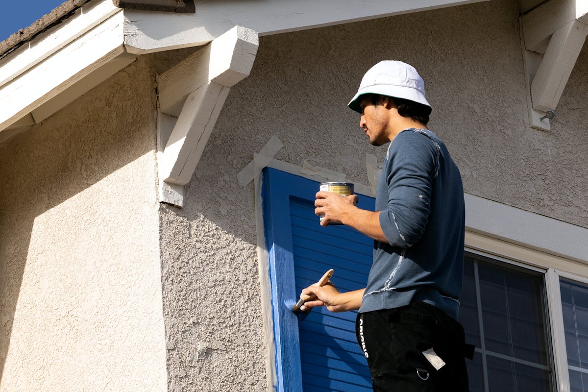 Man painting blue shutters on a stucco house while wearing a bucket hat.