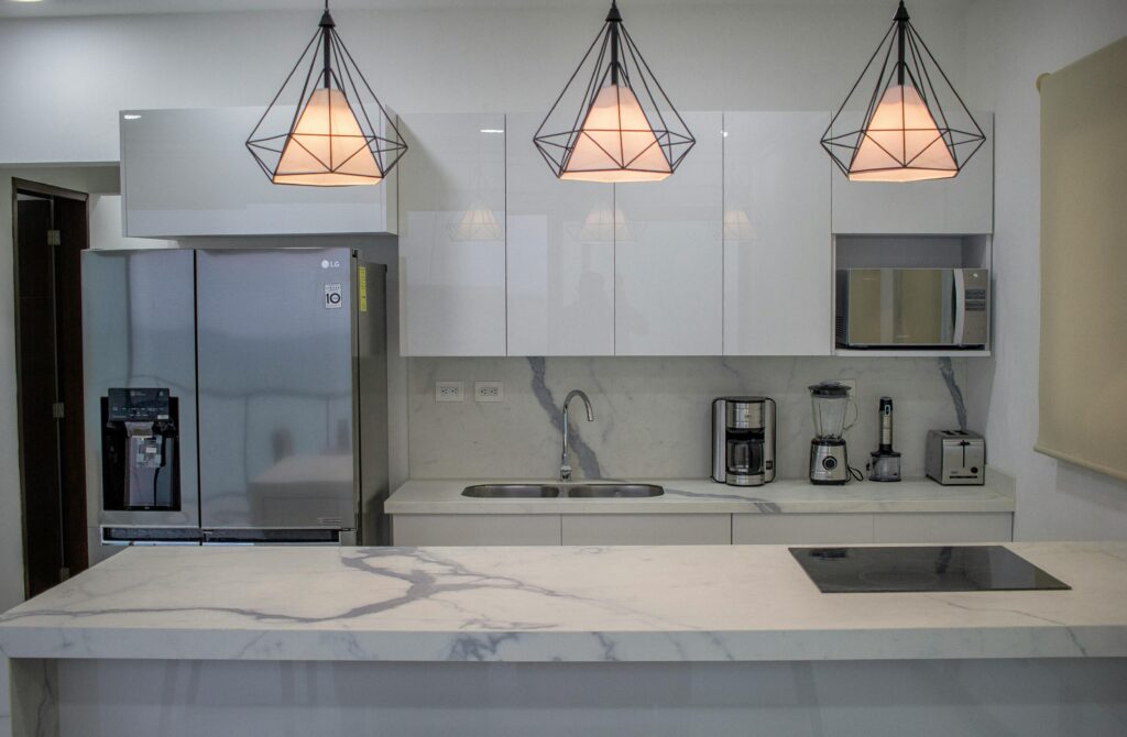 Modern white kitchen with marble worktop, pendant lights and stainless steel fridge.